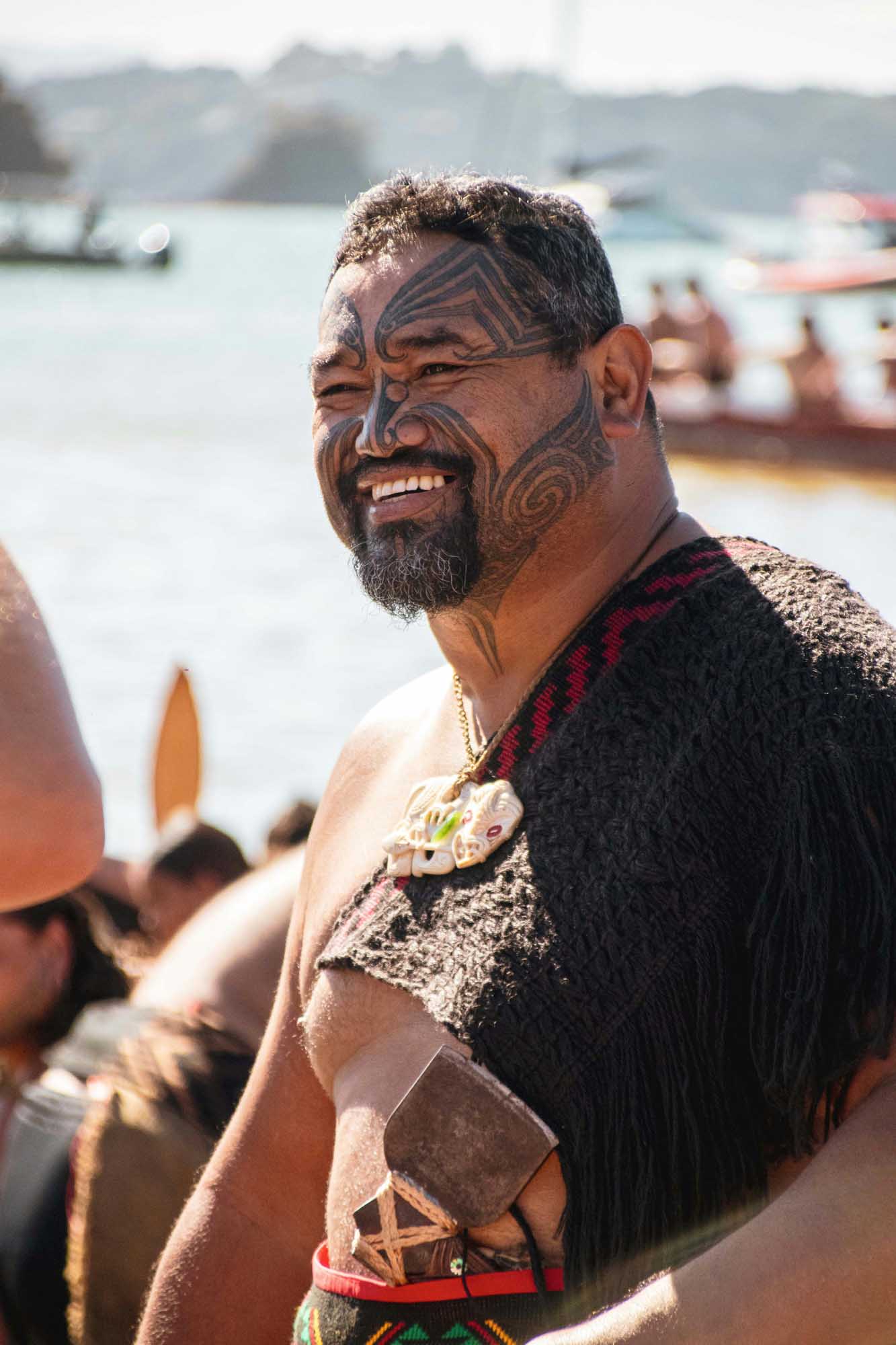 A man with traditional Maori ta moko facial tattoos smiles at the camera while standing near a waterfront with boats and hills in the background.
