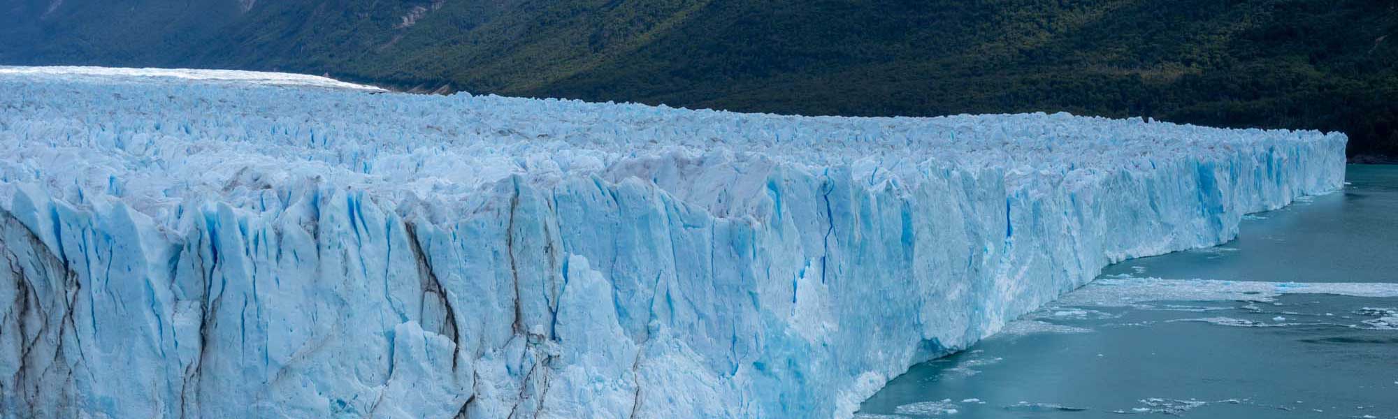 A dramatic view of a massive glacier terminus with striking blue-white ice formations rising from turquoise glacial meltwater, backed by dense forested mountains typical of Patagonia's stunning alpine landscape.