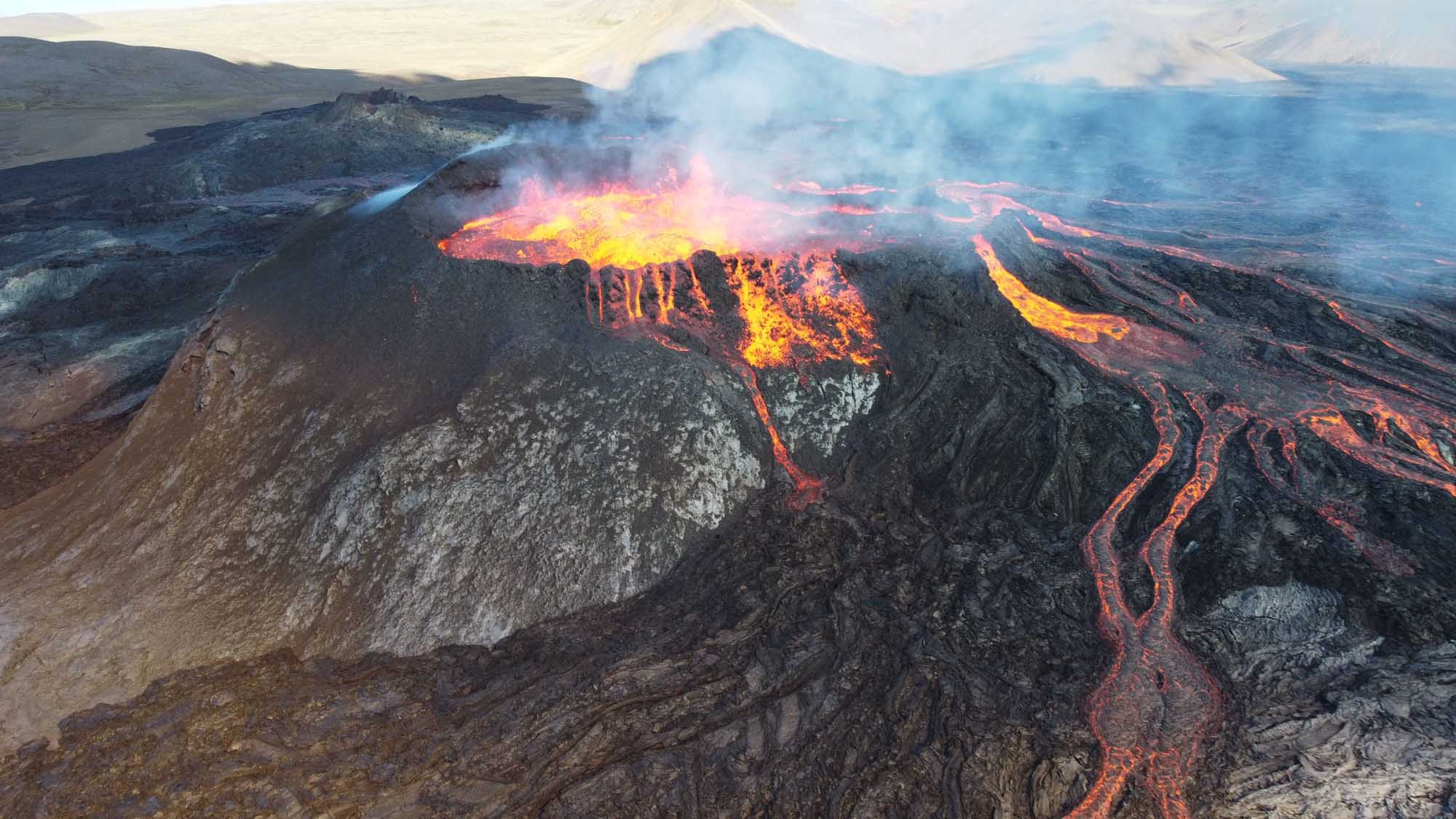 A dramatic aerial view of Mauna Loa volcano in Hawaii during an active eruption, with glowing lava fountains and flowing streams of molten rock cascading down the volcanic slopes, surrounded by dense volcanic rock formations and plumes of volcanic gases rising into the sky.