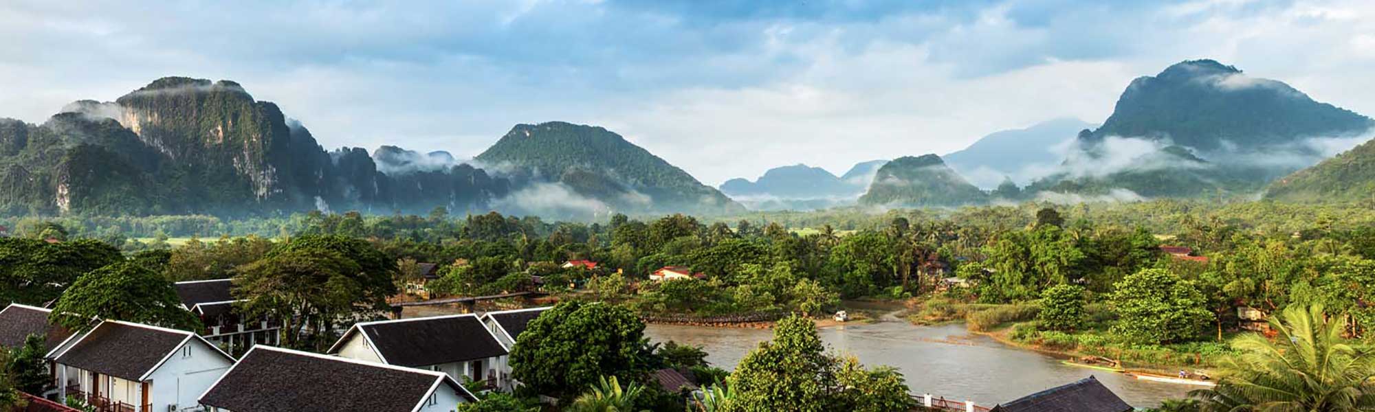 A scenic panoramic view of the Mekong River valley in Laos, featuring dramatic limestone karst mountains shrouded in morning mist, traditional riverside buildings with dark tile roofs, lush tropical vegetation, and the calm waters of the Mekong River in the foreground.