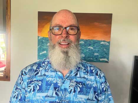 A smiling man with a white beard and glasses wearing a blue tropical Hawaiian shirt with palm tree and boat patterns, photographed indoors with a sunset seascape painting visible on the wall behind him.