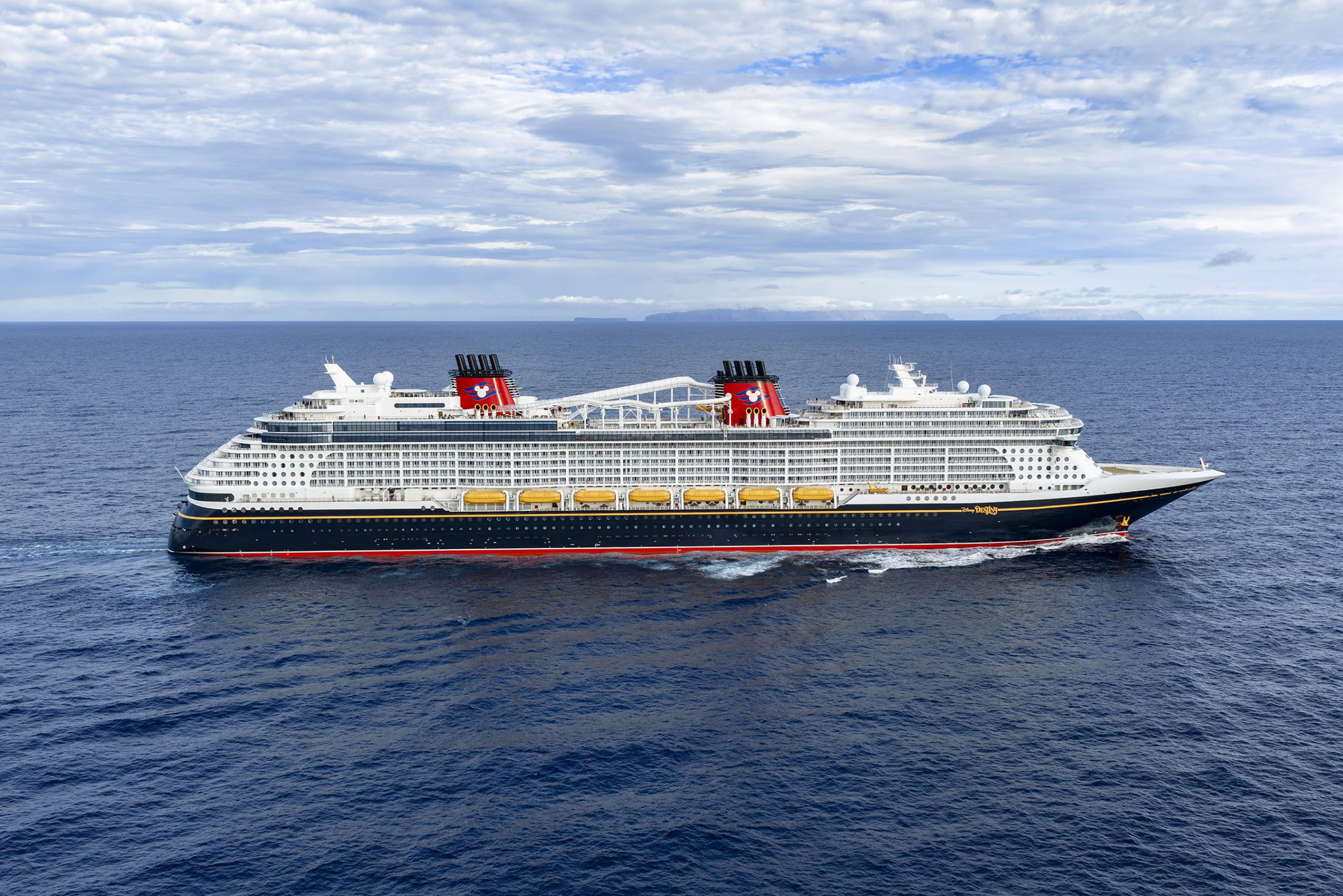 A large modern cruise ship with distinctive red funnels sails through calm blue waters under a partly cloudy sky with mountainous terrain visible on the horizon.