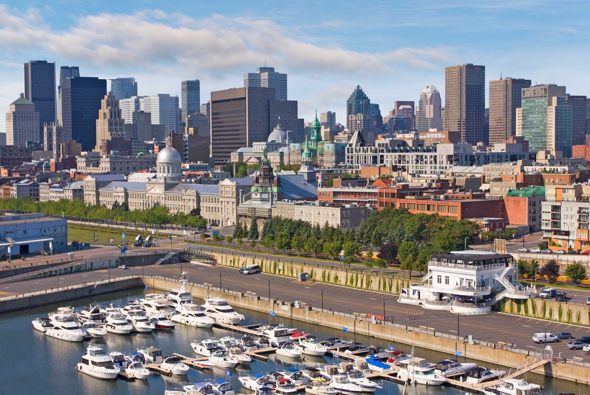 An aerial view of Montreal's downtown skyline featuring iconic skyscrapers and historic architecture, with a recreational marina filled with white yachts and boats in the foreground along the St. Lawrence River waterfront.
