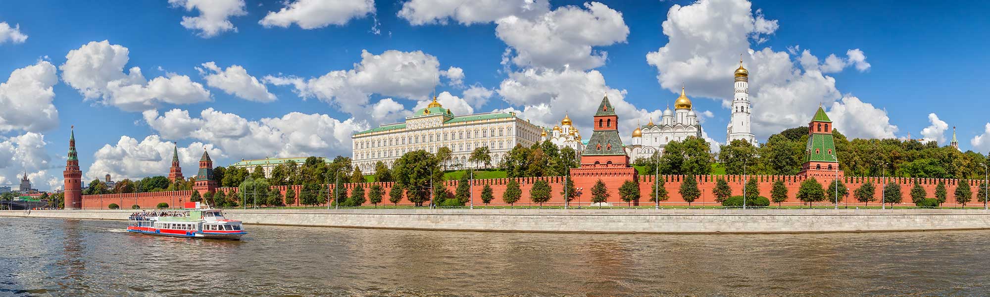 A scenic view of the iconic Moscow Kremlin fortress complex along the Moskva River, featuring its distinctive red brick walls, golden-domed cathedrals, and government buildings. A colorful tour riverboat passes in the foreground under clear skies with white clouds.