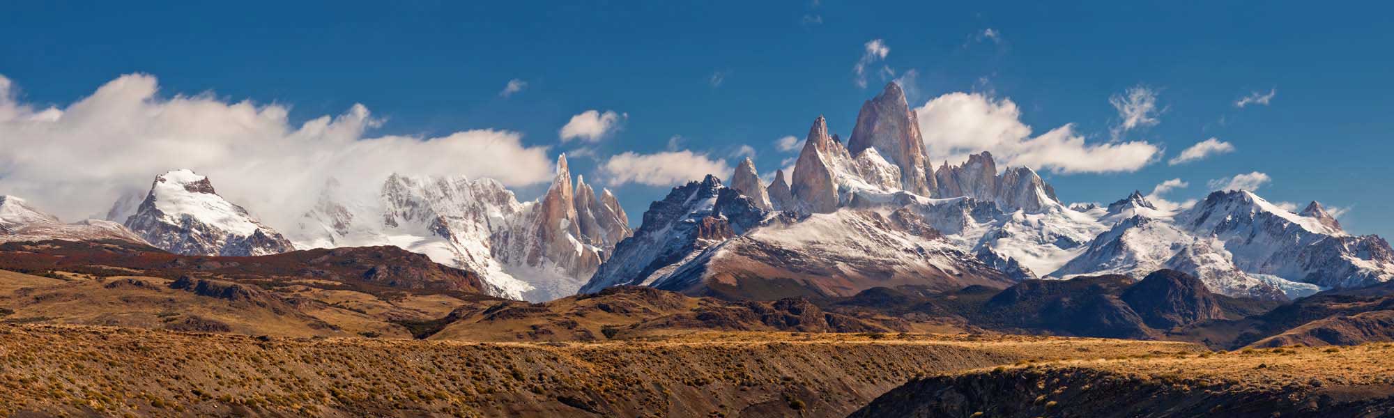 A dramatic panoramic vista of Mount Fitz Roy and the surrounding Patagonian mountain range in Argentina, featuring distinctive granite spires snow-capped peaks rising above arid, golden-brown steppe landscape under a brilliant blue sky with scattered white clouds.