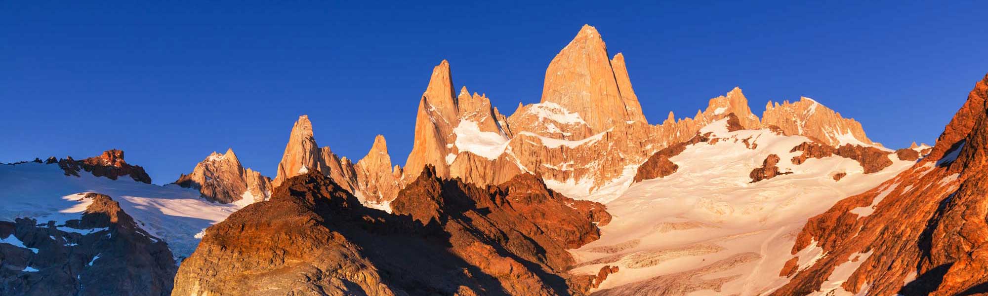 The iconic granite peaks of Mount Fitz Roy rise dramatically against a brilliant blue sky, their distinctive spires illuminated by golden morning light. Snow-covered glaciers and scree slopes surround the mountain in this stunning view of one of South America's most spectacular natural landmarks.