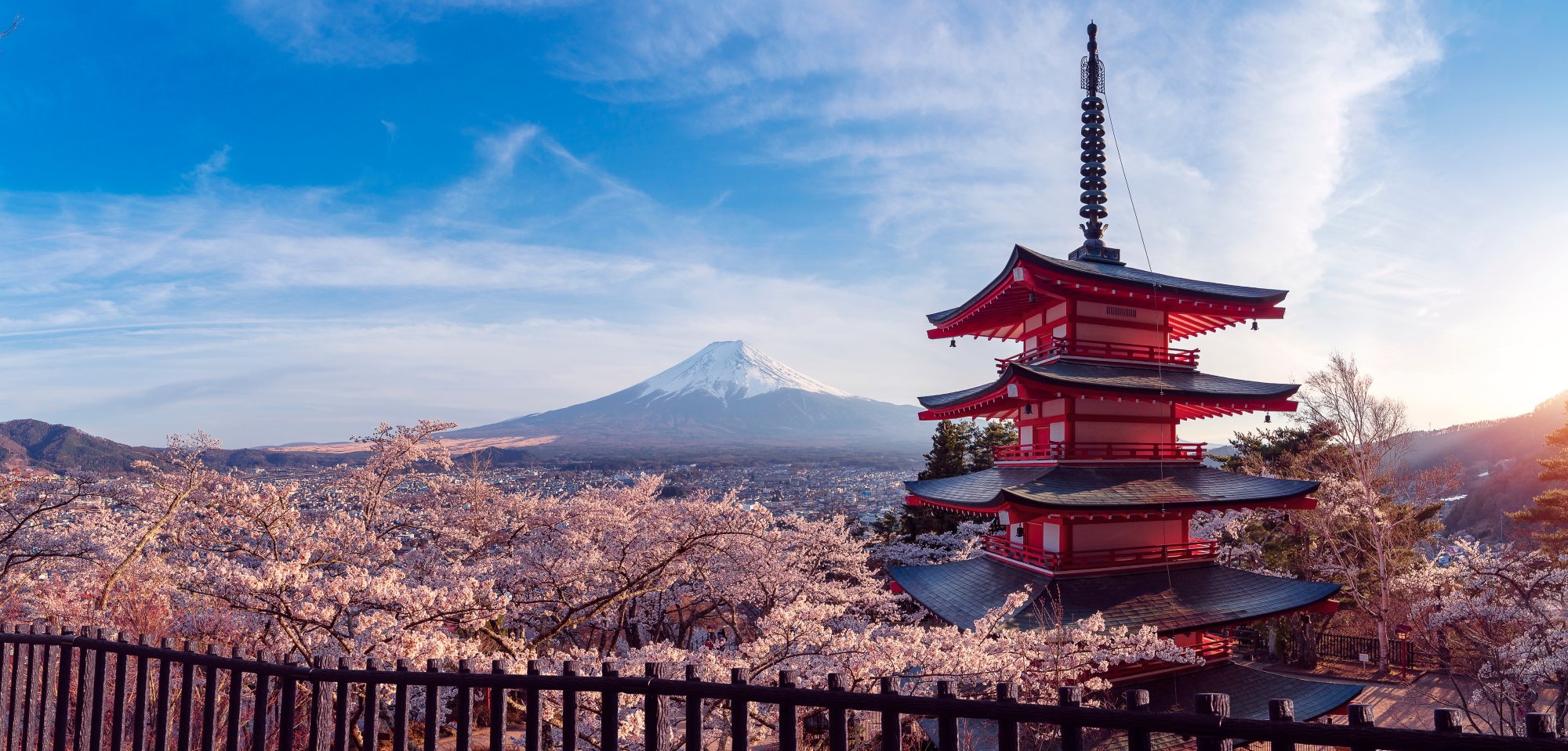 A stunning scenic view of a traditional red Japanese pagoda framed by cherry blossom trees in full bloom, with snow-capped Mount Fuji visible in the distance across the landscape.