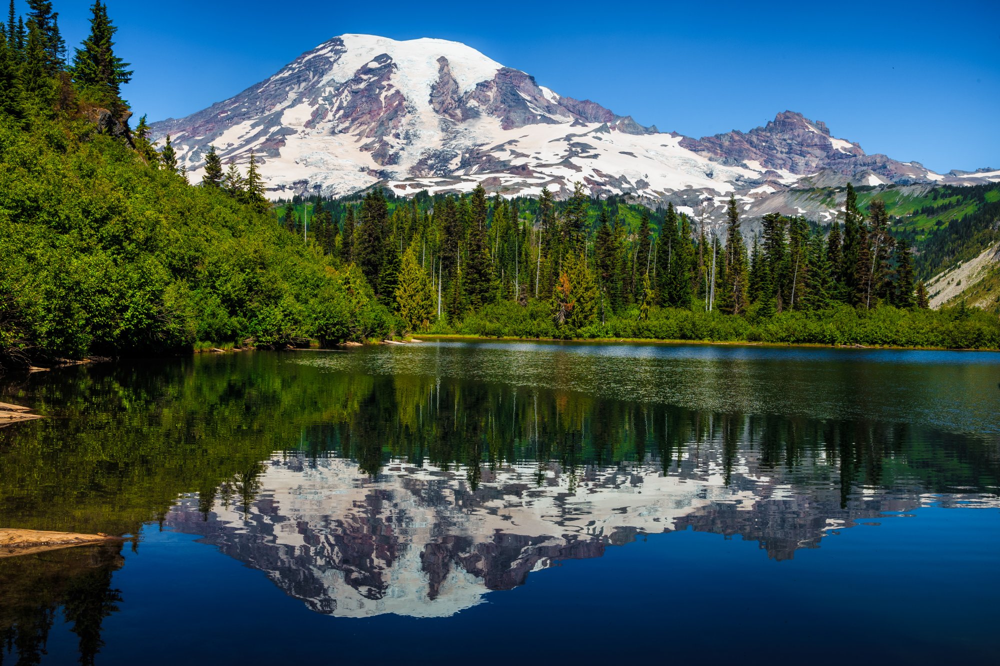 A stunning alpine landscape featuring Mount Rainier's snow-capped summit perfectly reflected in a pristine mountain lake, surrounded by dense coniferous forest under clear blue skies.
