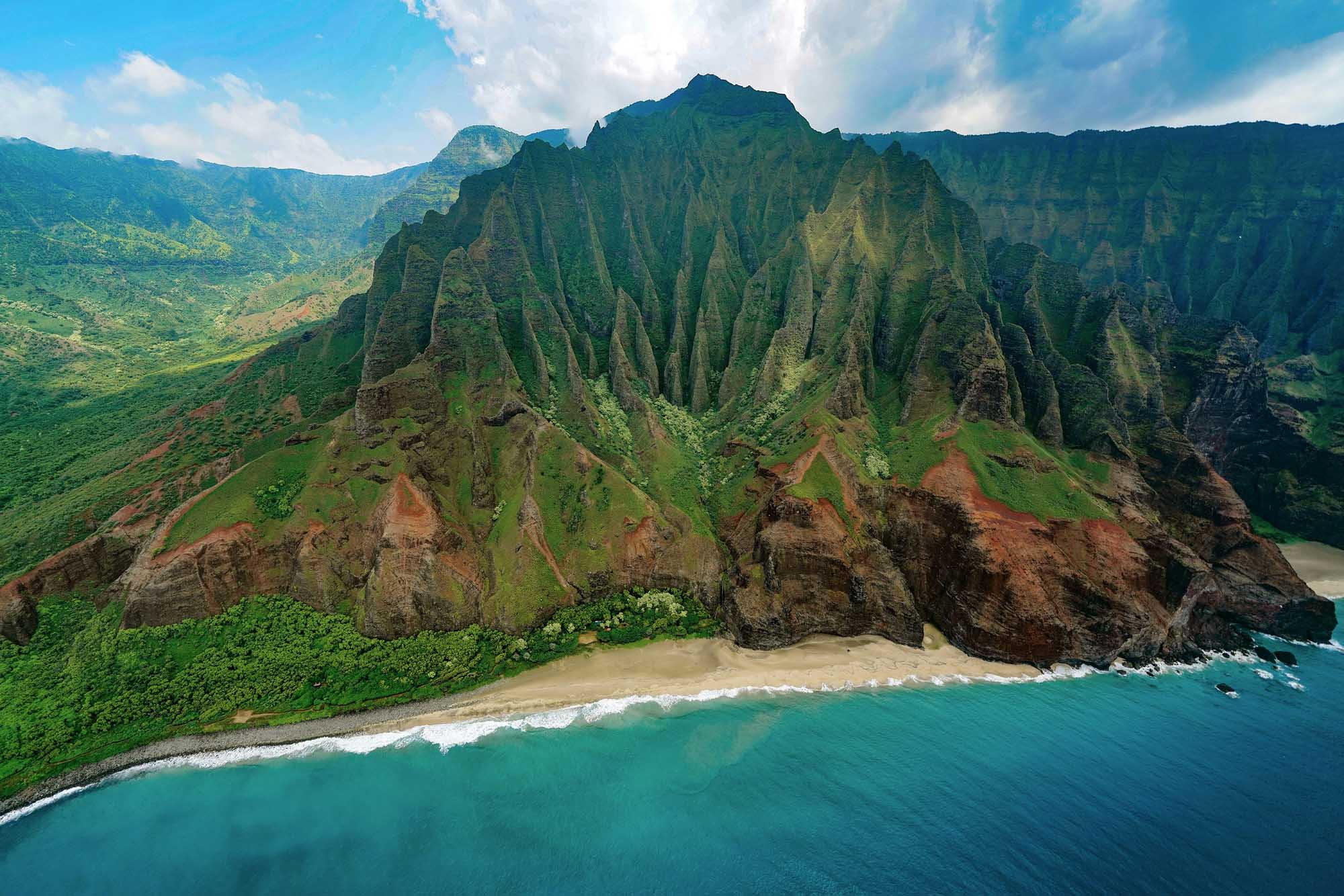 A stunning aerial view of the iconic Na Pali Coast featuring dramatic emerald-green fluted sea cliffs rising vertically from a pristine sandy beach and turquoise Pacific Ocean waters. The distinctive sharp ridges and valleys carved into the mountainous terrain create a striking natural landscape characteristic of this UNESCO World Heritage site.