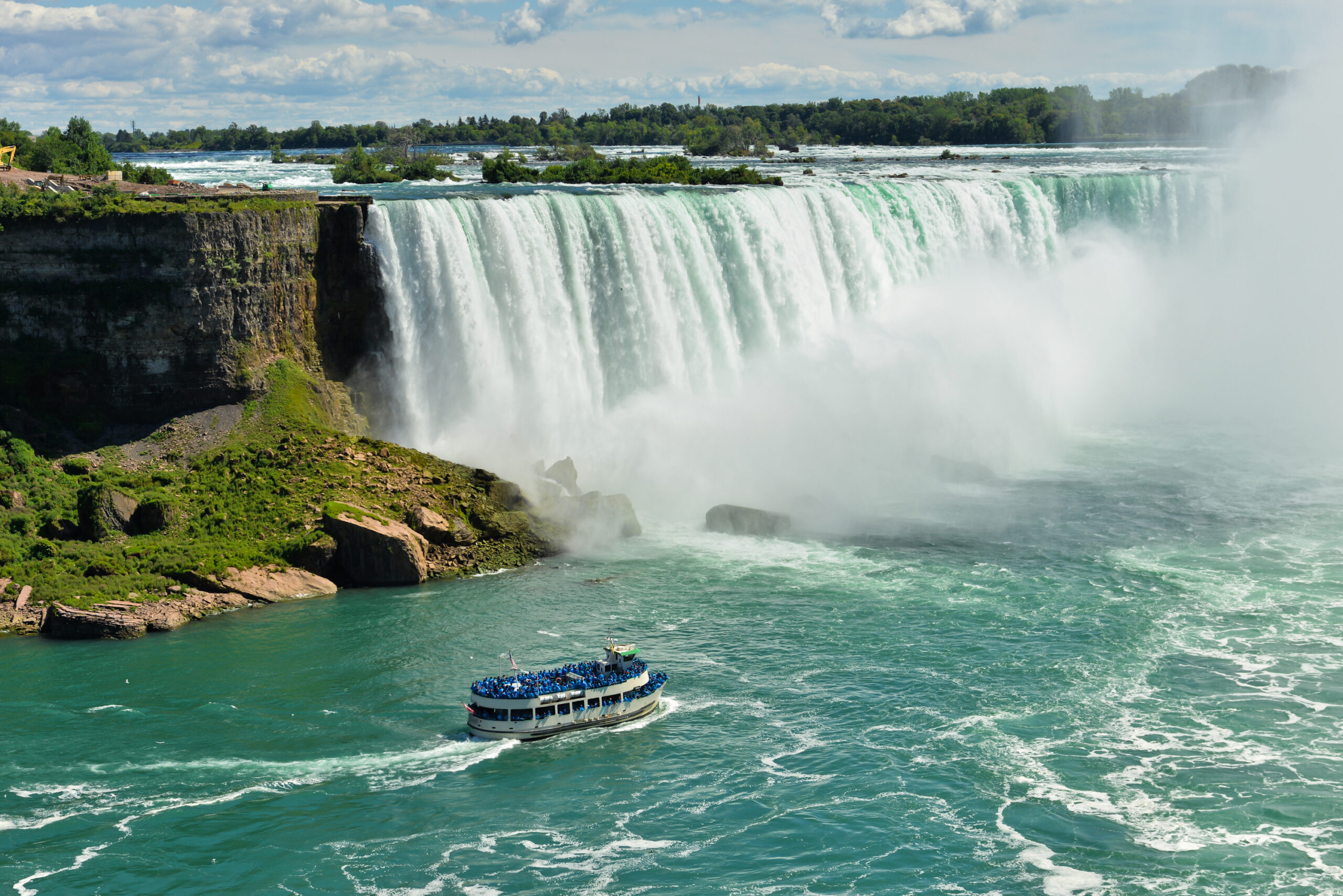 A scenic view of Niagara Falls Horseshoe Falls with a passenger tour boat navigating the turquoise waters below, surrounded by mist and lush greenery on the Canadian side. The iconic natural wonder showcases the massive cascade of water with a blue-sky backdrop and forested landscape.