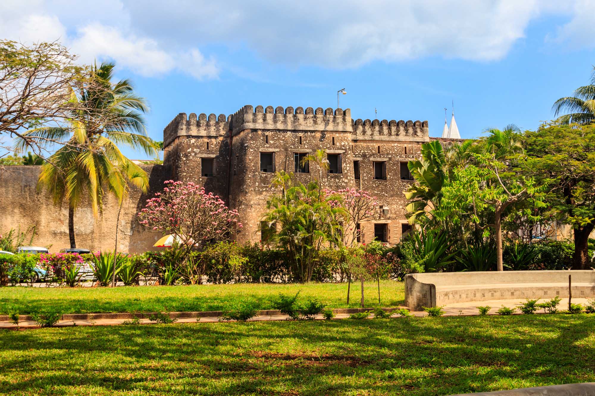 The historic Old Fort (Arab Fort), a centuries-old stone fortification in Stone Town, Zanzibar, is surrounded by lush tropical gardens with blooming pink flowers and palm trees. The massive crenellated walls and multiple windows showcase classic Arab-Swahili architecture, with a flag flying from the rooftop against a clear blue sky.