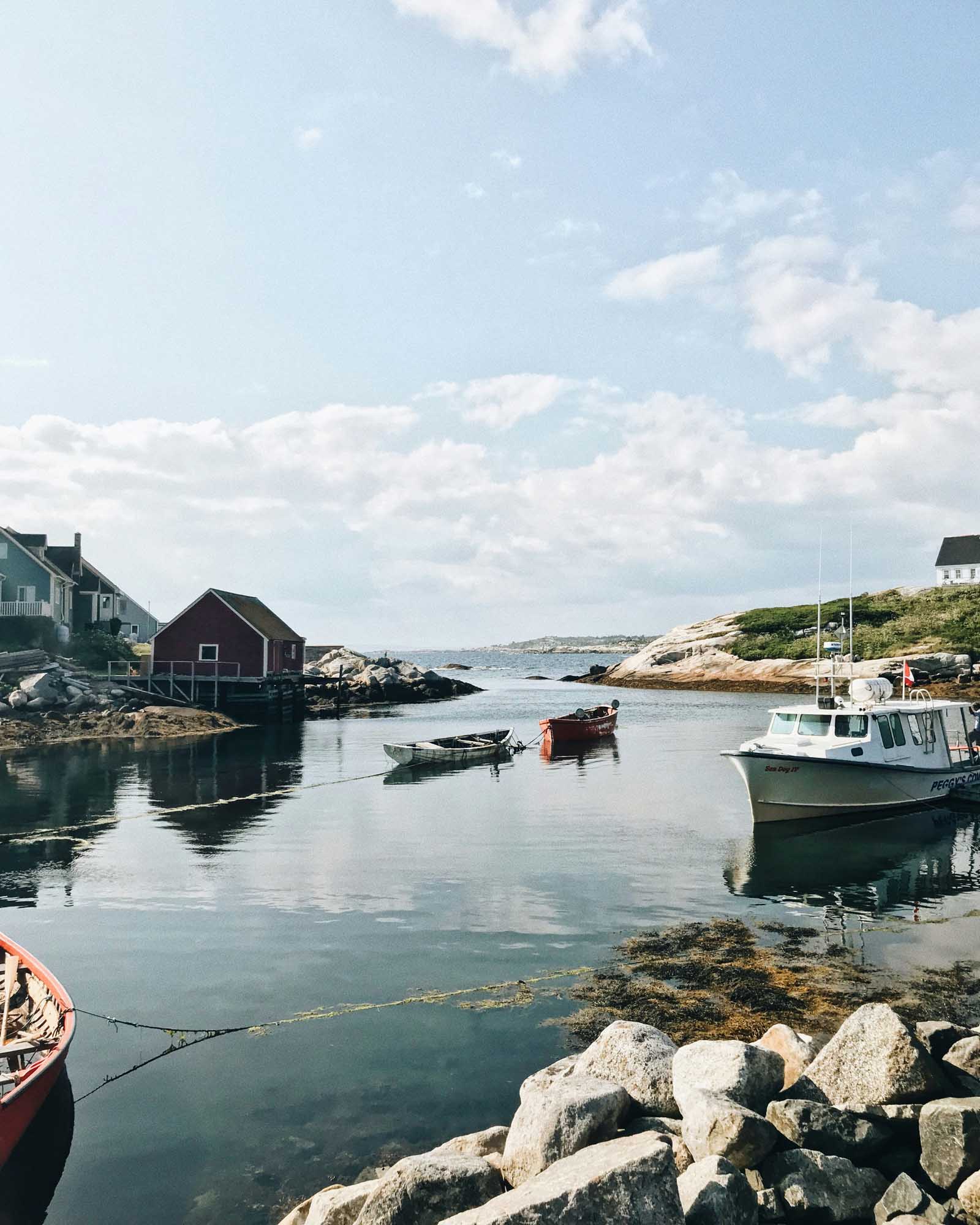 A serene coastal fishing village with traditional wooden buildings, moored fishing boats, and rocky shoreline under a partly cloudy sky.