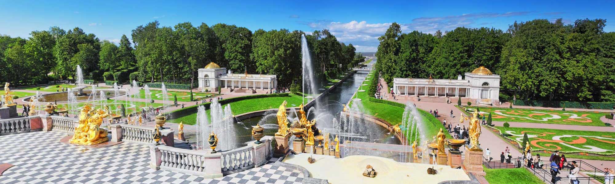 An aerial view of the magnificent Peterhof Palace complex featuring elaborate golden sculptural fountains, geometric formal gardens, cascading water features, and neoclassical pavilions with golden domes, surrounded by verdant parkland and tree-lined avenues in Russia.