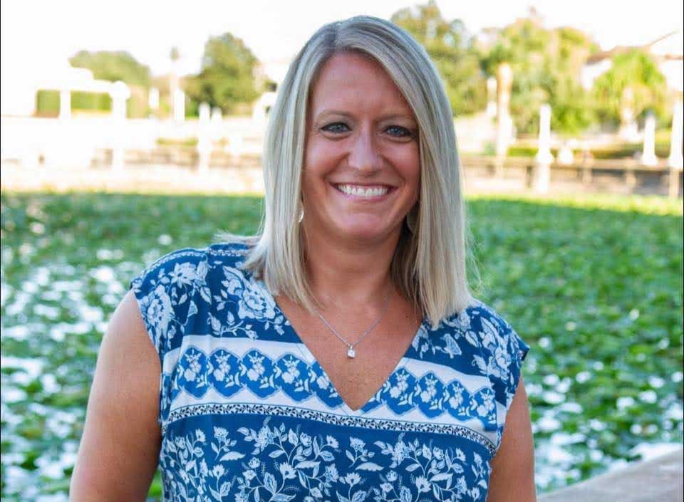 A smiling woman with blonde hair wearing a blue and white patterned sleeveless top poses for a portrait in front of an agricultural field with blooming crops and farm structures visible in the background.