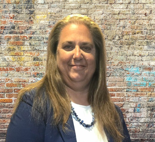 A professional headshot of a woman with long brown hair wearing a dark navy blazer over a white top, posed against a textured brick wall background with weathered red, orange, and blue paint.