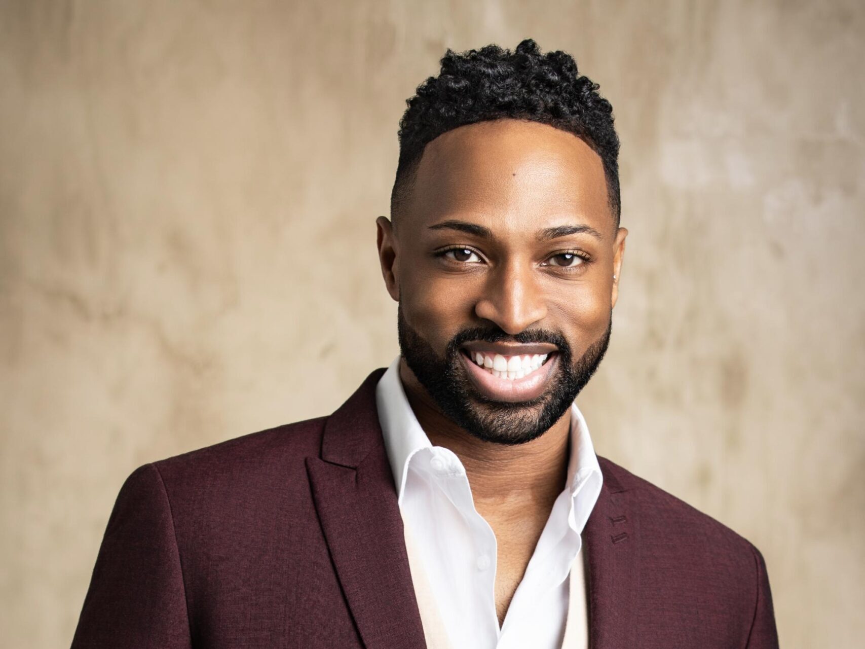 A professional headshot of a man wearing a burgundy blazer over a white shirt, smiling at the camera against a neutral beige background. This is a polished corporate or business portrait style photograph.