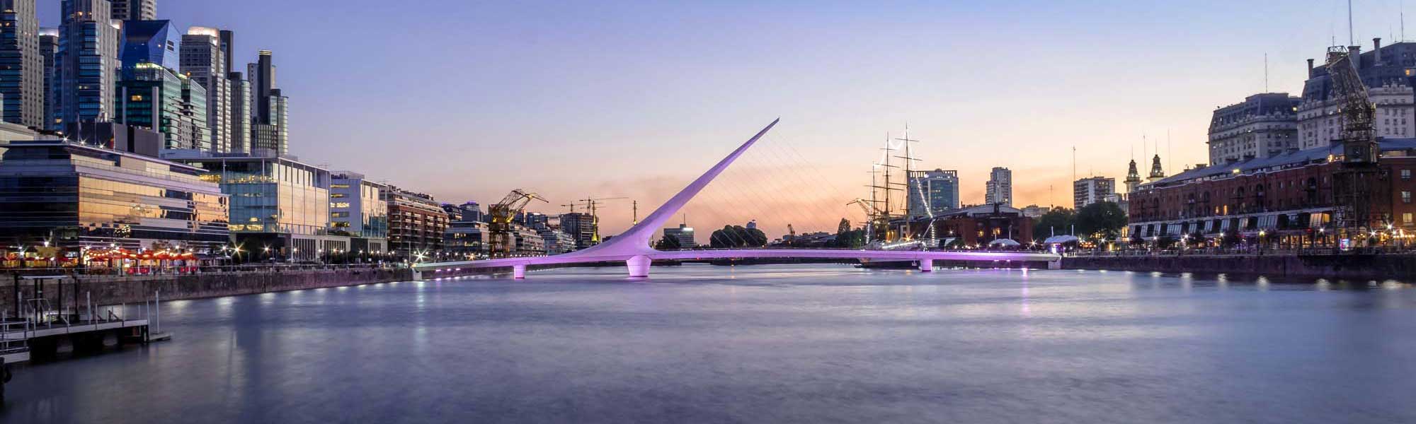 A stunning twilight view of the iconic Puente de la Mujer (Woman's Bridge) illuminated in purple lights, spanning a calm waterway in the Puerto Madero neighborhood of Buenos Aires. The modern cable-stayed bridge is framed by contemporary architecture, historic buildings, and tall ships, creating a striking contrast between old and new urban development.