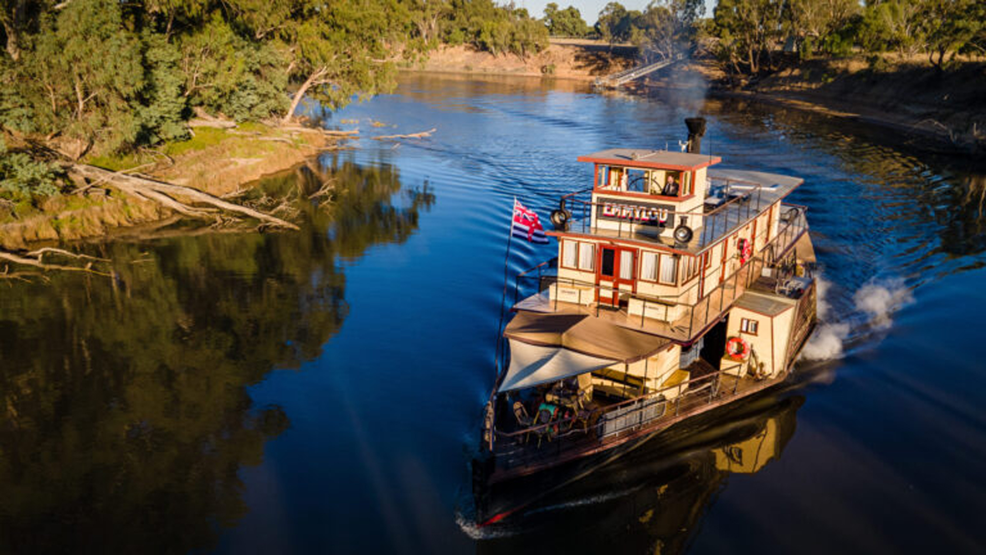 A multi-deck paddle steamer or riverboat navigates a serene inland river lined with native eucalyptus trees and lush vegetation. The vessel features distinctive orange and cream-colored superstructure with multiple decks, railings, and a flag, representing a classic Australian river cruise experience in what appears to be a regional waterway.