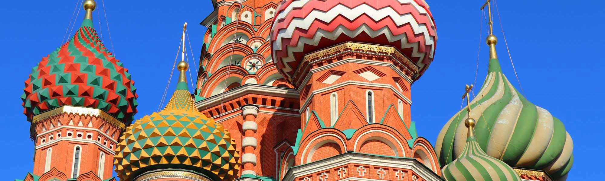 A striking Russian Orthodox church featuring distinctive colorful onion domes with intricate geometric patterns in red, green, gold, and white against a clear blue sky. The ornate architecture showcases traditional Russian ecclesiastical design with multiple spires and elaborate decorative elements characteristic of Eastern Orthodox religious buildings.