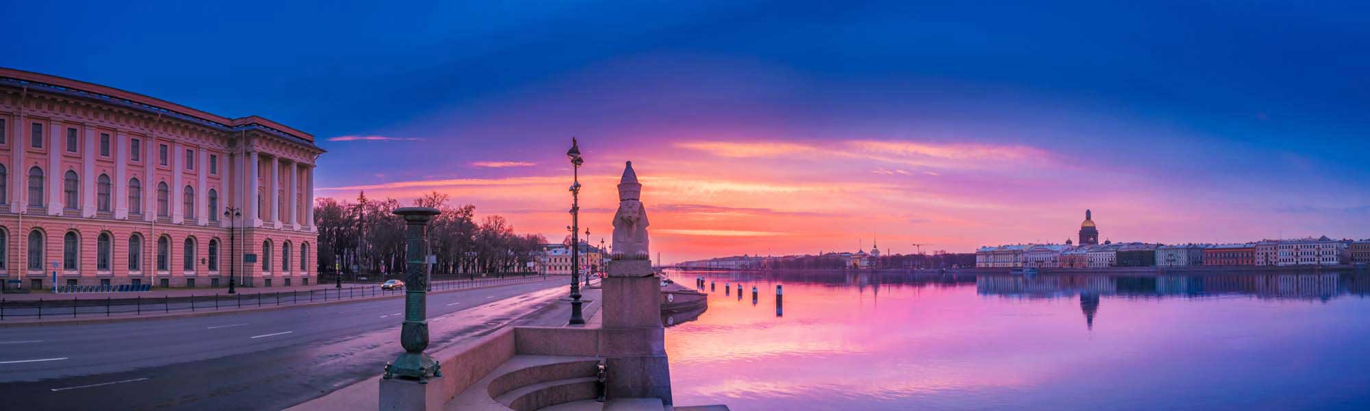 A breathtaking dawn scene over the Neva River in Saint Petersburg, Russia, featuring the iconic pink neoclassical building on the left, ornate lamp posts, and the city's historic skyline reflected in the calm water under a vibrant purple and orange sunrise sky.