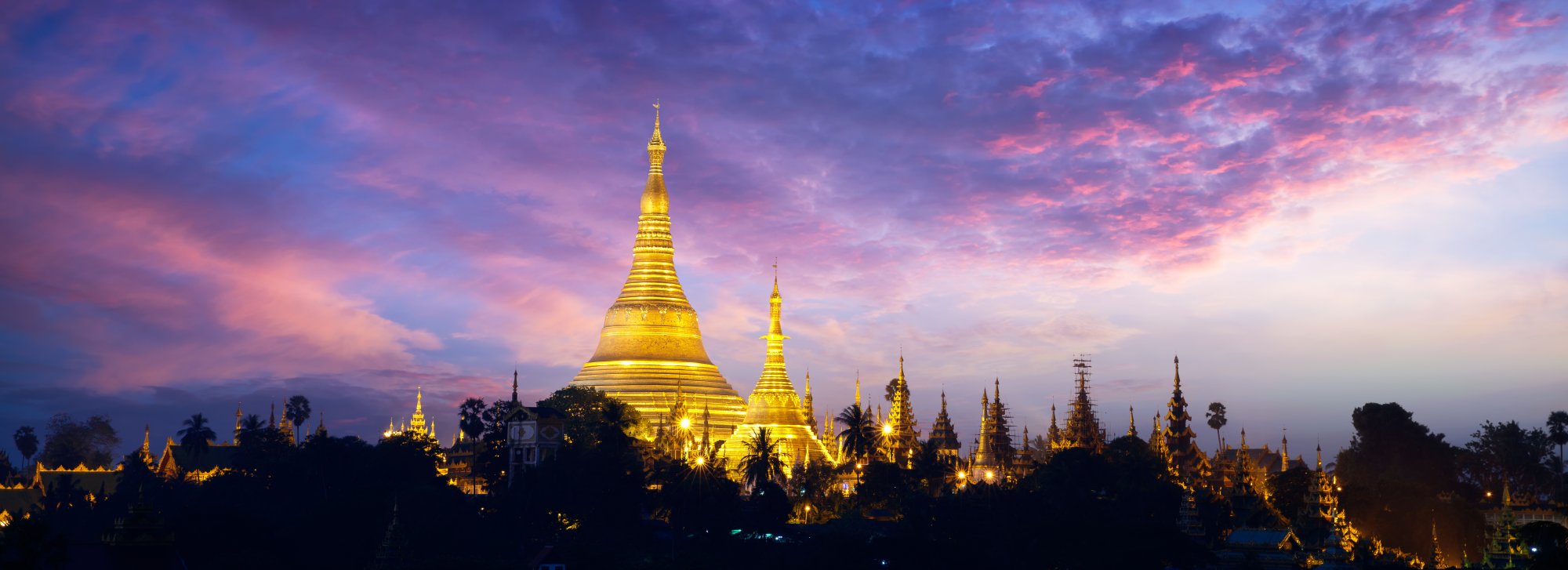 The iconic golden Shwedagon Pagoda illuminated against a dramatic purple and pink twilight sky, with its distinctive tiered spire rising prominently above the surrounding temple complex and palm trees. The sacred Buddhist monument glows warmly in the evening light, showcasing the intricate architecture of Myanmar's most revered spiritual landmark.