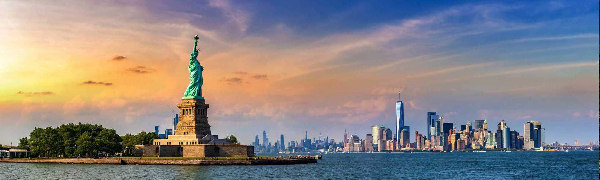 An iconic view of the Statue of Liberty on Liberty Island with the Manhattan skyline illuminated in golden sunlight during sunset. The image captures the copper statue against a vibrant sky with the modern skyscrapers of lower Manhattan visible across the water.