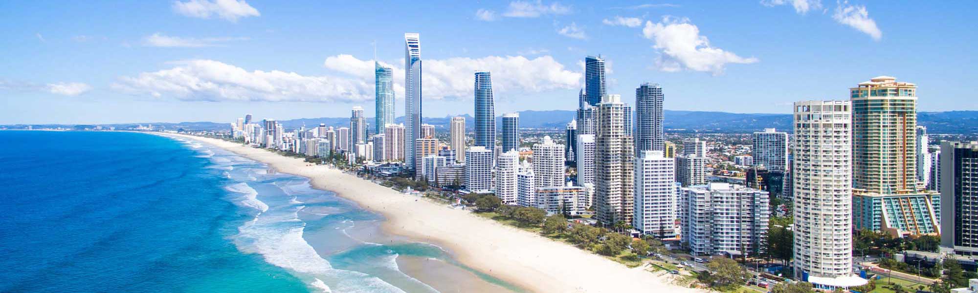 An aerial view of Australia's Gold Coast featuring a stunning high-rise beachfront skyline with modern condominiums and hotels lining pristine golden sand beaches adjacent to sparkling turquoise ocean waters with white-capped waves.