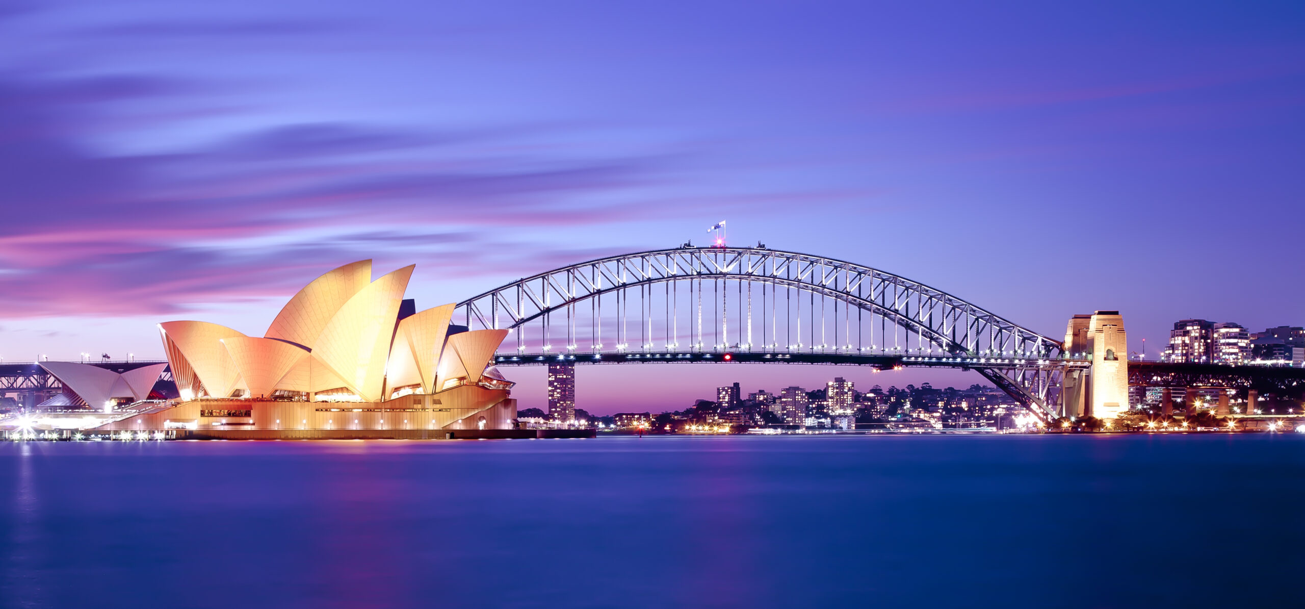 An iconic twilight photograph of Sydney's most famous landmarks, featuring the illuminated Sydney Opera House in the foreground and the Sydney Harbour Bridge spanning across the harbor against a stunning purple and pink sunset sky, with the city's skyline lights twinkling in the background.