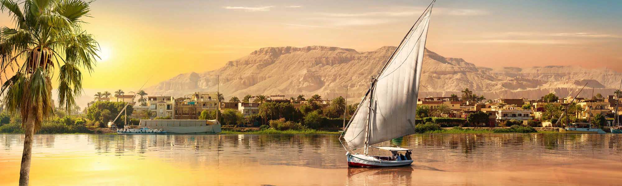 A traditional Egyptian felucca sailboat with white canvas sails glides peacefully across the Nile River at golden sunrise, with palm-lined riverbanks, colorful buildings, and desert cliffs in the background.