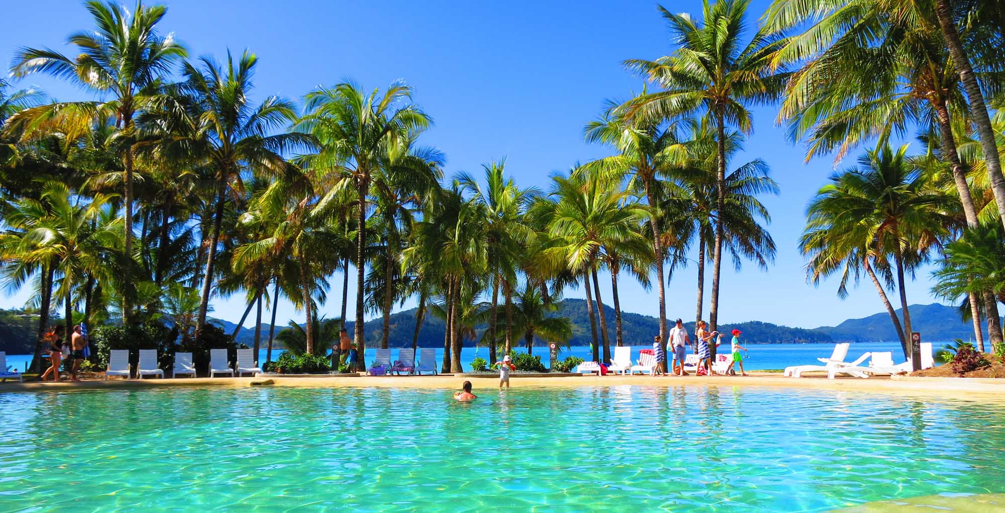 A stunning tropical beach scene featuring crystal-clear turquoise waters, pristine white sand, and numerous swaying palm trees. Visitors relax on lounge chairs while others enjoy swimming in the idyllic lagoon, with forested mountains visible across the water under a clear blue sky.