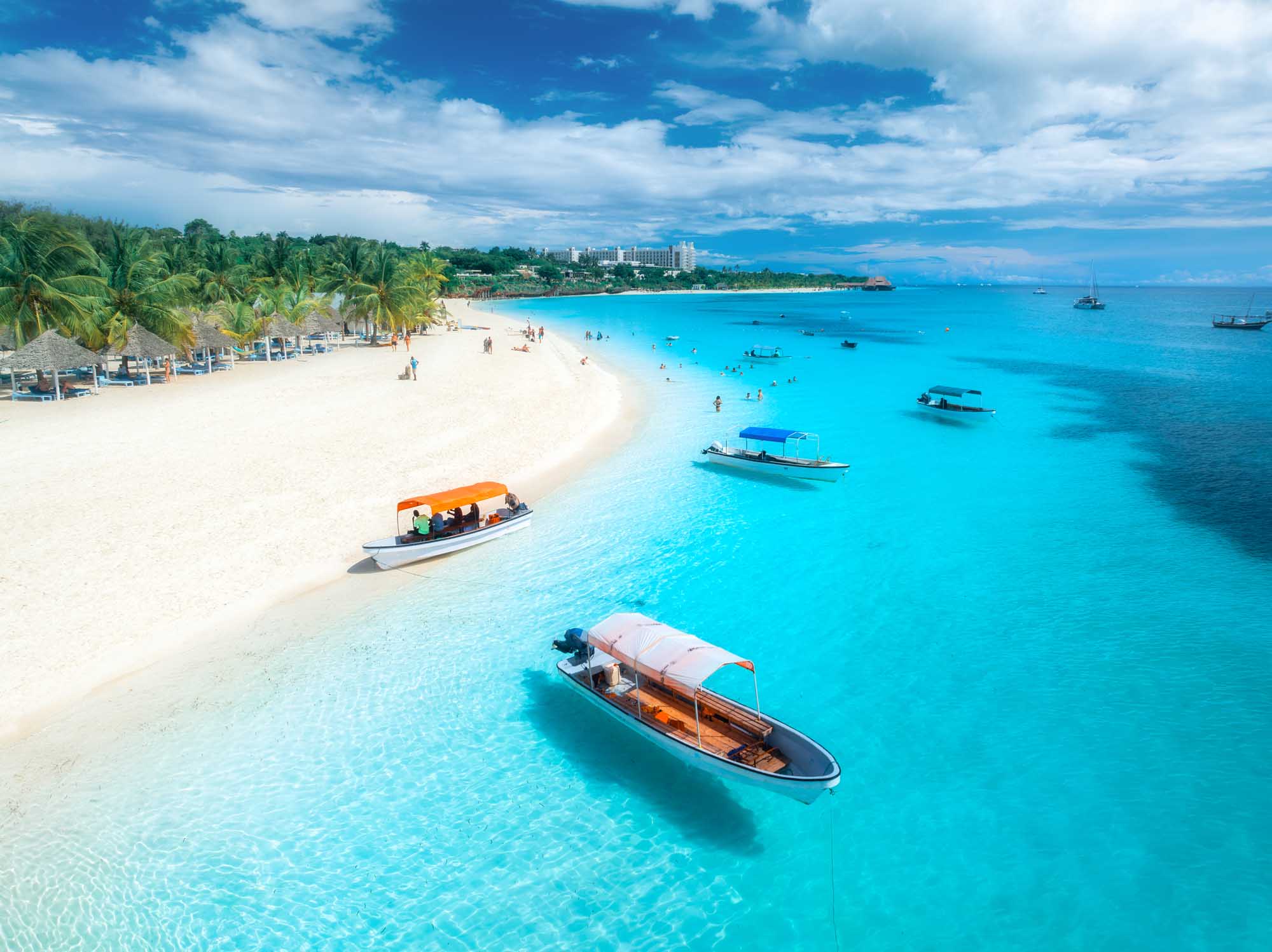 An aerial drone view of a pristine tropical beach featuring crystal-clear turquoise waters, white sand, traditional fishing boats moored offshore, palm trees lining the shore, and a beachfront resort in the background under a bright blue sky.