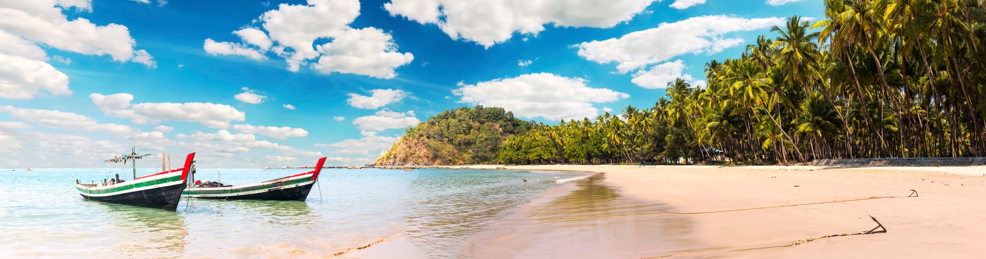 A picturesque tropical beach scene featuring two traditional colorful long-tail boats anchored in crystal-clear turquoise waters, with pristine white sand, lush palm trees, and a forested hillside under a brilliant blue sky with white clouds.