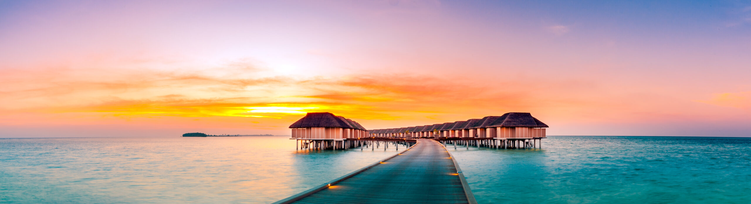 A serene tropical resort featuring traditional thatched-roof overwater bungalows connected by wooden walkways, set in crystal-clear turquoise waters during a spectacular golden sunset with warm orange and pink sky reflections.