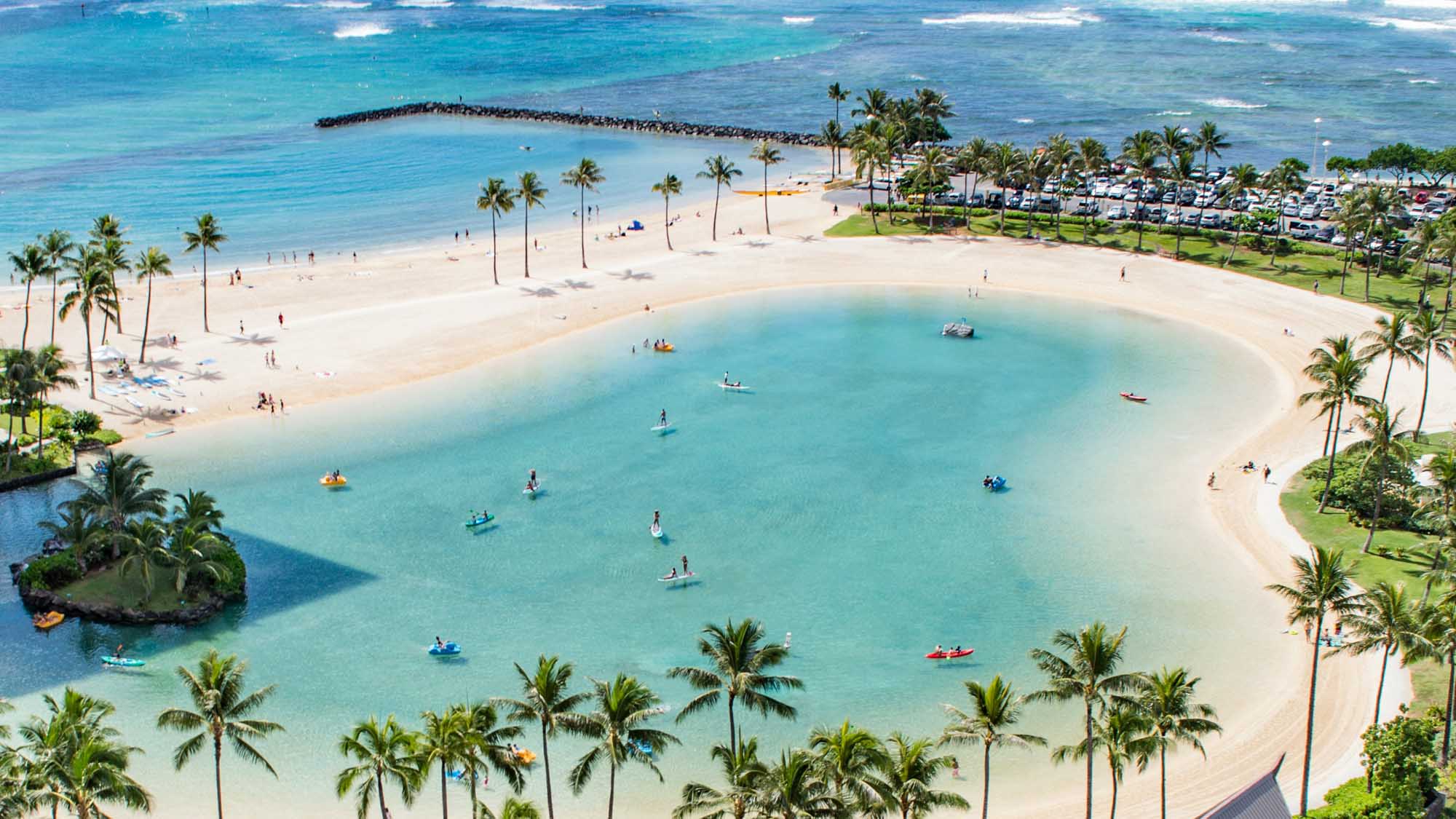 An aerial view of a pristine tropical beach destination featuring a stunning turquoise lagoon with soft white sand, scattered palm trees, and numerous water activities. The scene includes a parking area, grassy park space, and a protective breakwater, showcasing a popular beach resort destination with calm, crystal-clear waters ideal for swimming and water sports.