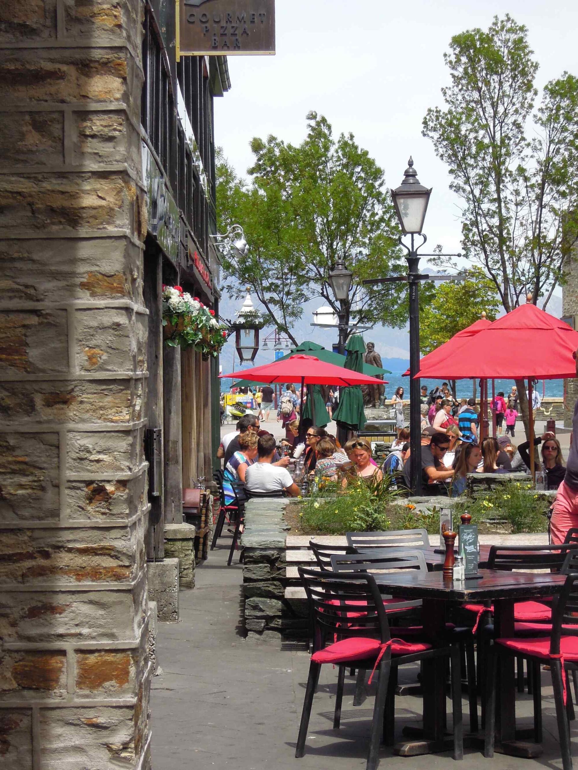 A bustling waterfront dining area features black tables with red cushioned chairs under bright red umbrellas, with a stone building featuring a gourmet bar sign on the left. In the background, a scenic beach with mountains is visible, populated with beachgoers and additional red umbrellas creating a vibrant summer atmosphere.