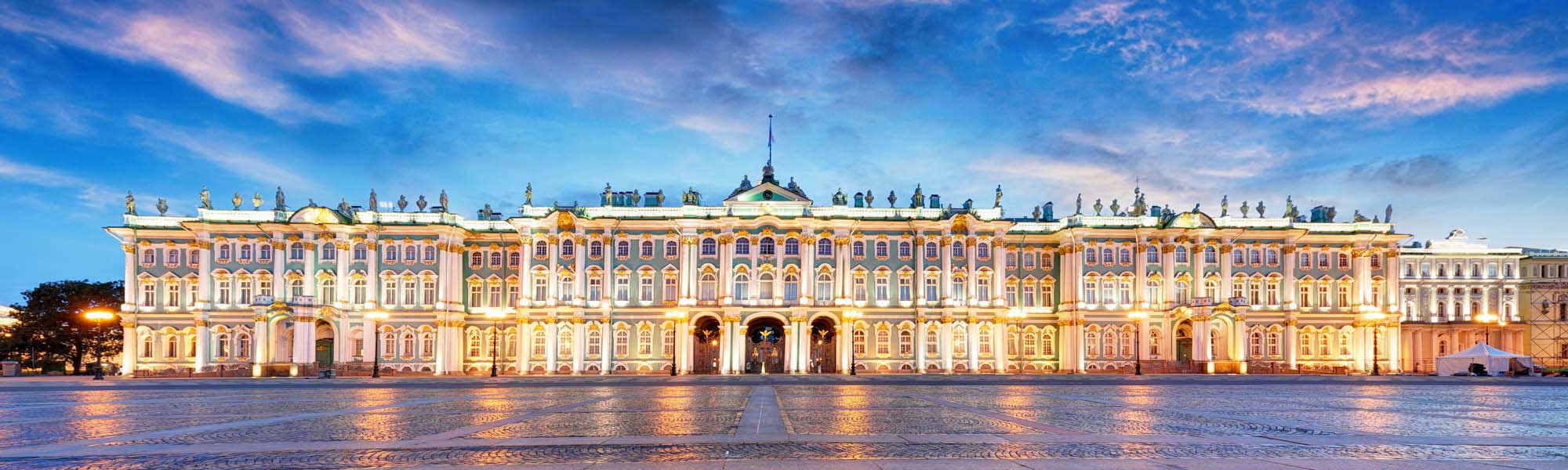 The iconic Winter Palace illuminated at dusk, showcasing its distinctive pink and white Baroque architectural facade across Palace Square in Saint Petersburg, Russia. The grand 18th-century imperial residence reflects warm golden light on the wet plaza surface beneath a blue twilight sky.