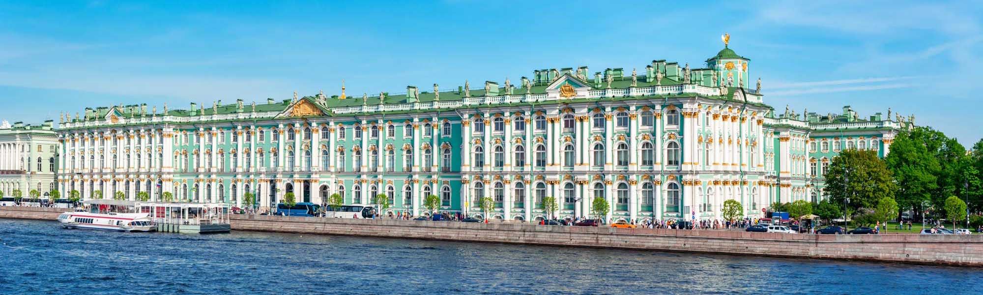 The magnificent Winter Palace in St. Petersburg, Russia, displaying its iconic turquoise and white Baroque facade with golden ornamental details, as viewed from the Neva River. The grand 18th-century imperial residence features multiple stories of arched windows, decorative moldings, and a distinctive green roof punctuated by ornamental spires.