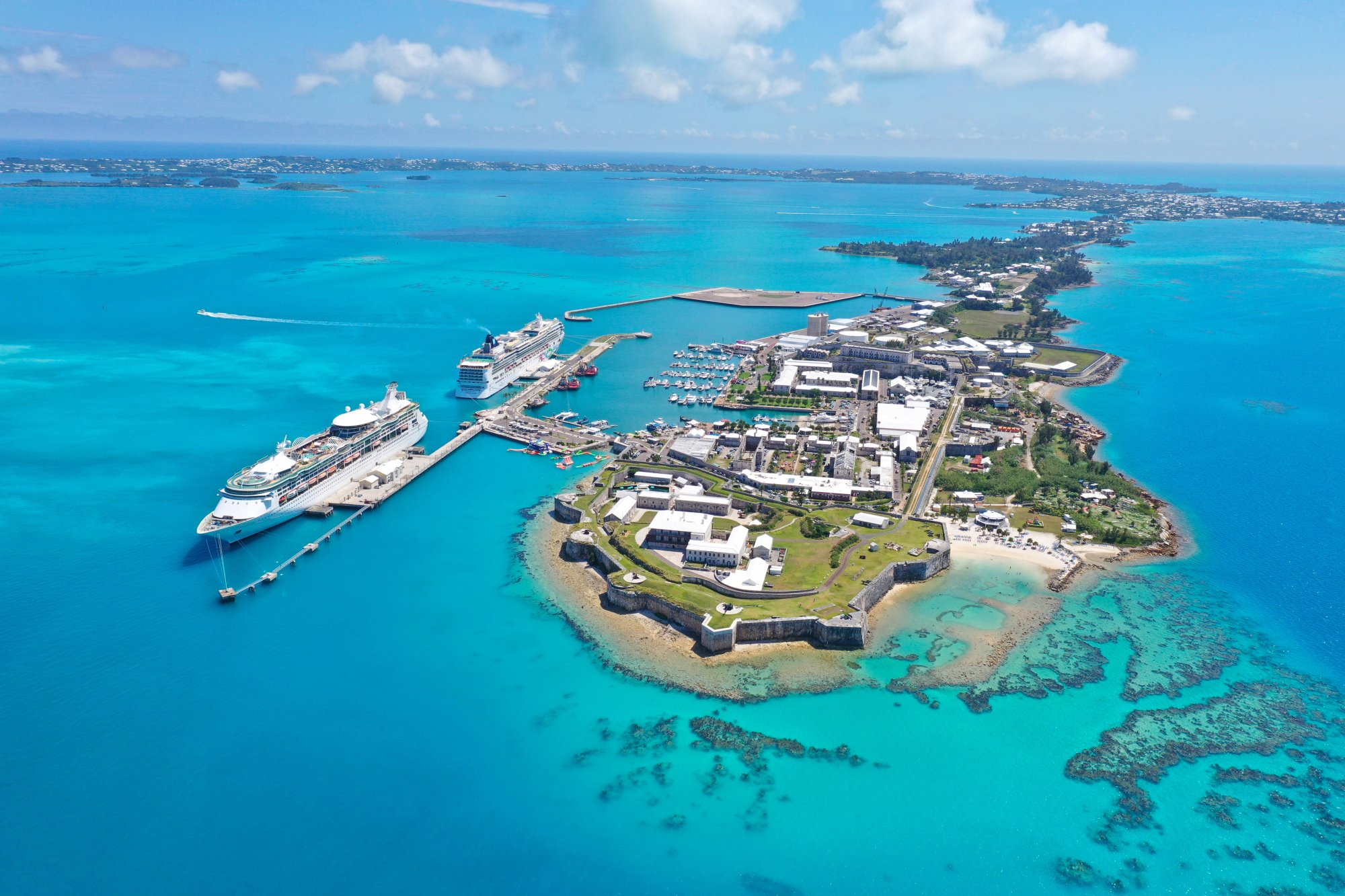 An aerial view of a large cruise ship docked at King's Wharf in Bermuda, showcasing the iconic fortress and colonial architecture of the island alongside turquoise waters.