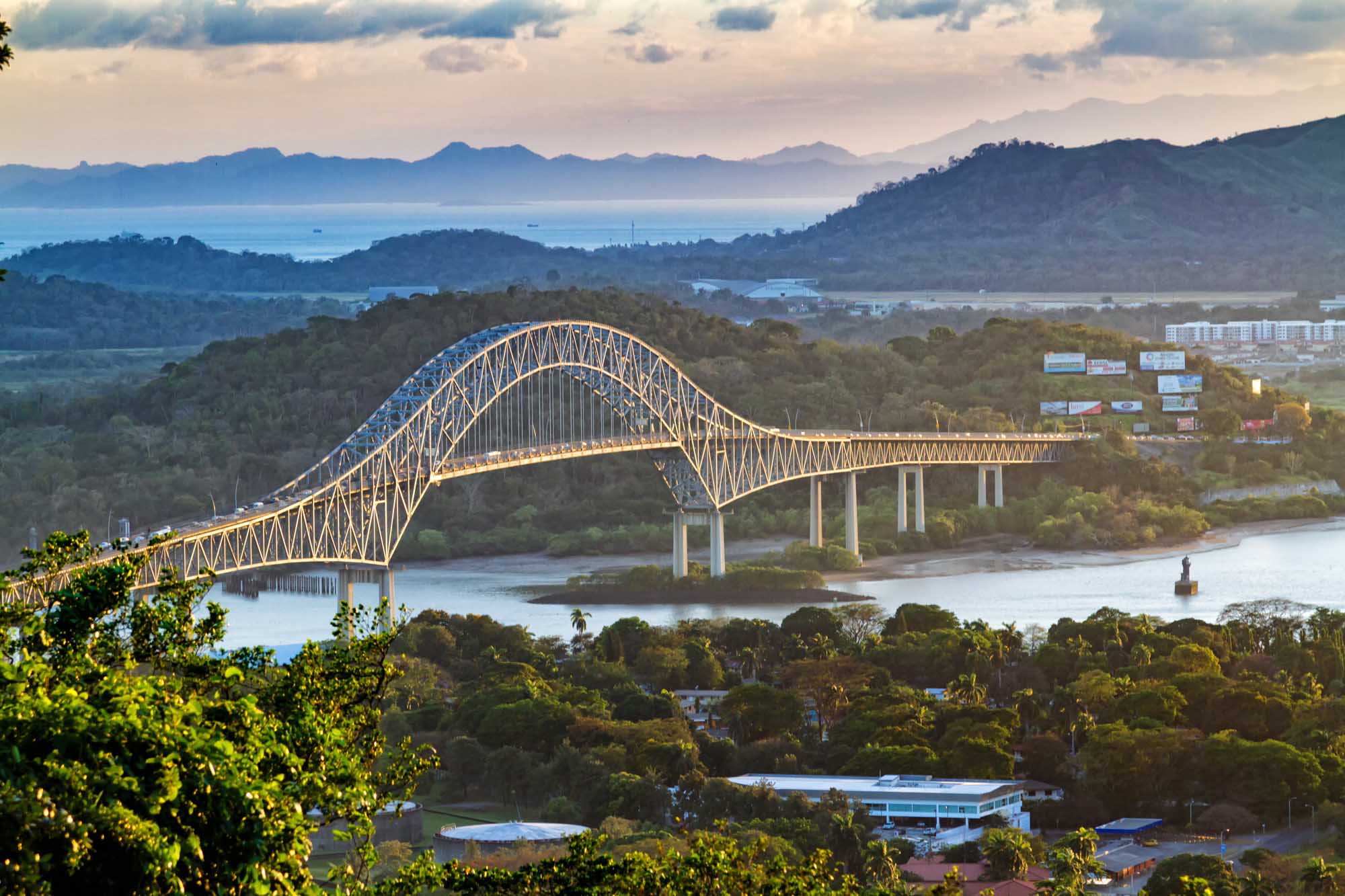 A stunning arched bridge with golden-yellow steel lattice construction spans a wide river in a lush, mountainous tropical landscape.