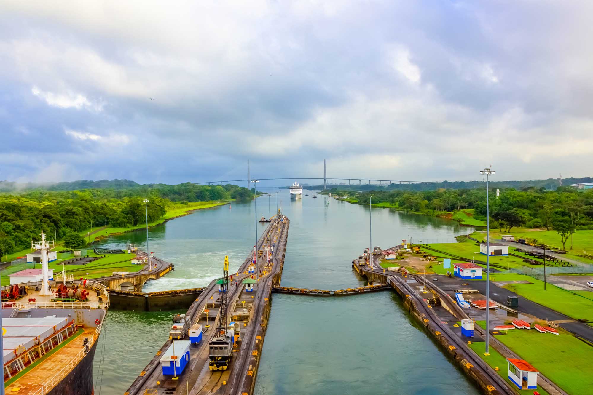 An aerial view of the Panama Canal's lock system showing massive lock chambers with ships in transit, with lush tropical landscape surrounding the infrastructure.