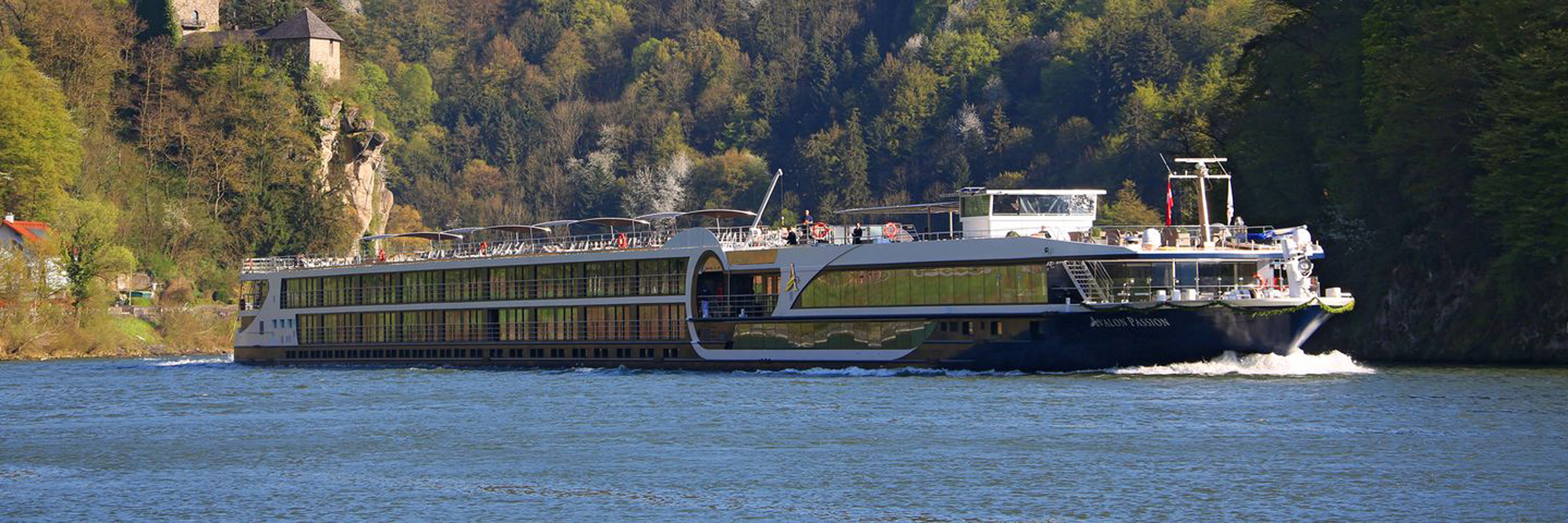 A modern river cruise ship navigates a scenic European river corridor flanked by forested cliffs